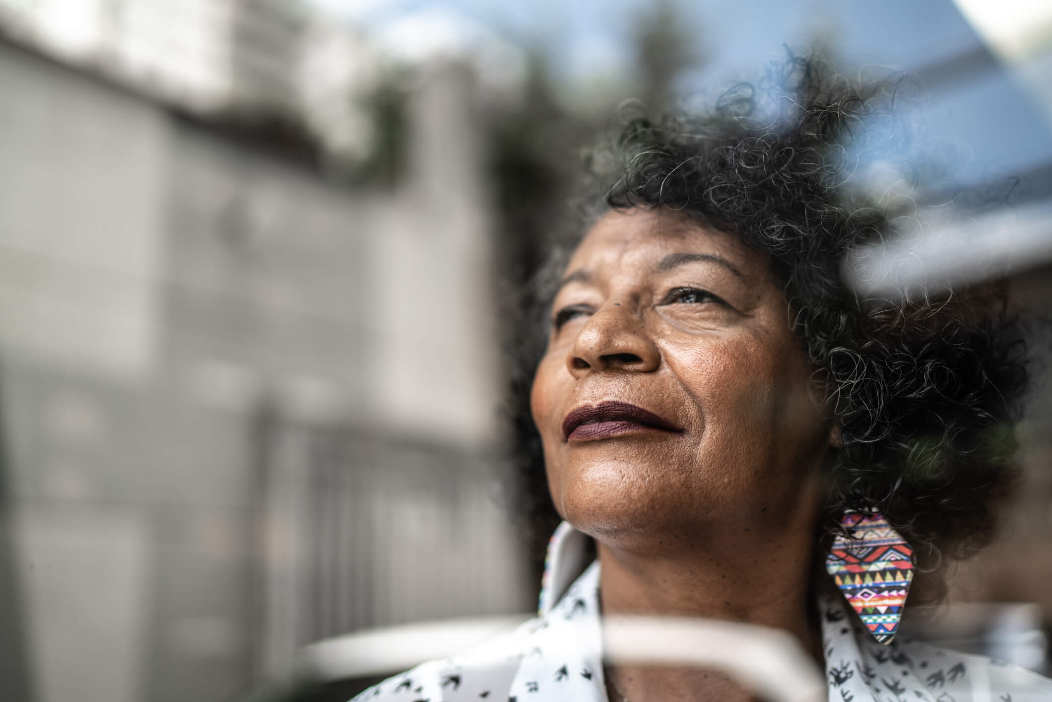 A dark-skinned, older woman pictured from the collarbone up looks away from the camera as if she is looking at something in the distance.